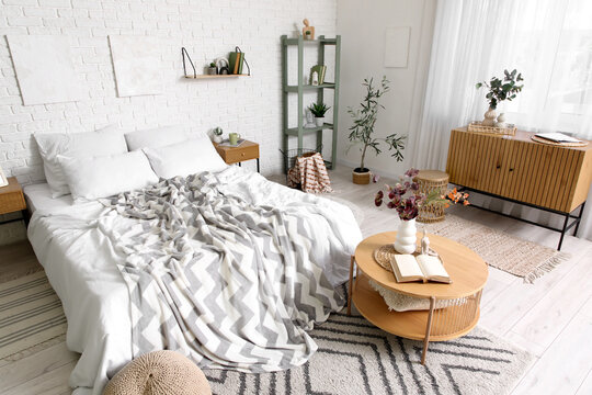 Interior of bedroom with big bed, shelving unit, bedside table, commode, coffee table and different beautiful artificial plants