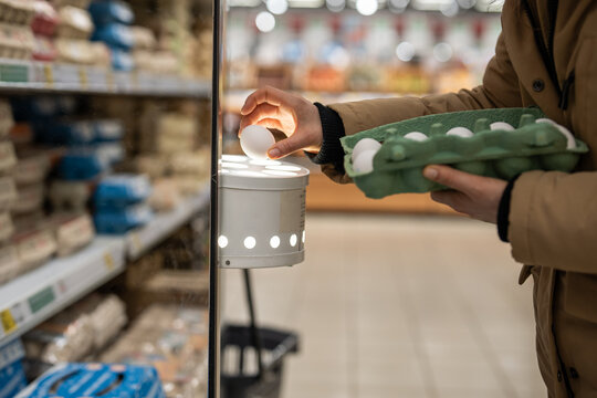 Unrecognizable woman in supermarket putting raw egg from box inside special ovoscope to check quality of eggs by candling light through shell. Person examinees condition of eggs in grocery store.