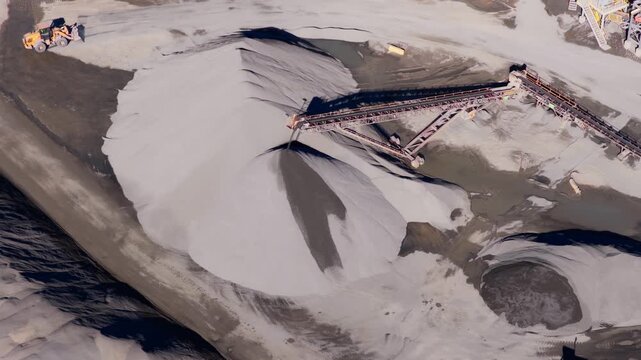 Top down aerial of conveyor belt casting shadow spreading over gravel pile as front loaders drive
