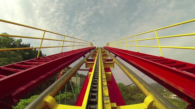 Point-of-view footage from the front of a roller coaster ascending toward the peak of the track. The bright red and yellow rails, chain lift mechanism, and safety railings are clearly visible.