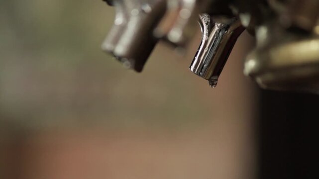 Close-up footage of a metallic espresso machine portafilter spout with a droplet of liquid hanging from its tip. The blurred background emphasizes the spout and the droplet, capturing a precise moment