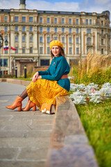 A woman in a stylish turquoise top and yellow skirt is smiling at the camera as she walks through the city. She is wearing a beret and carrying a small bag.