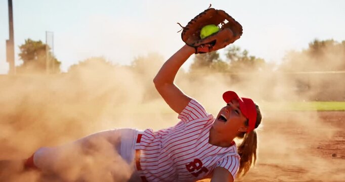 Young female athlete diving to catch a softball on a dusty field during a game at sunset