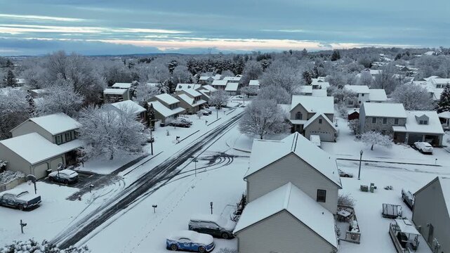 Cold morning over snowy suburban neighborhood with gabled houses and bare trees. Aerial view during sunrise at winter with snow in American housing development. Flyover drone shot.