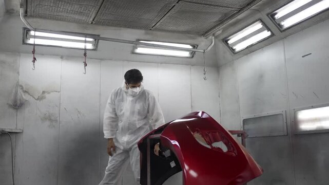 The mechanic is spraying a car bumper at a car repair shop. engineer, land vehicle, car repair shop, small business.