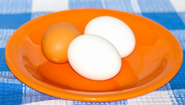 A vibrant orange plate holds three eggs arranged in a face pattern, with two white eggs for eyes and one brown egg for the nose. The plate sits on a blue and white plaid cloth.