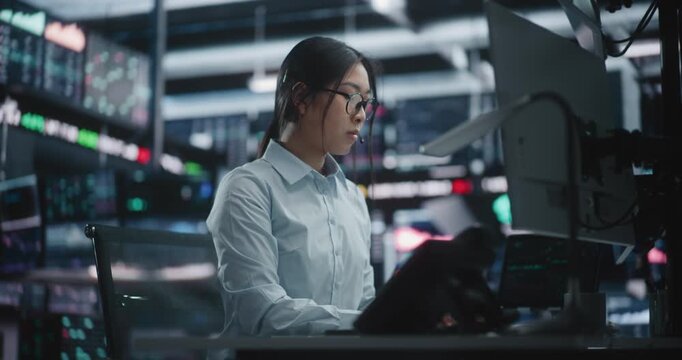 Female Trader Analyzing Real Time Stock Market Data at Large Screens. Focused Asian Woman in Glasses Works Intently at High Tech Desk, Typing on Keyboard While Monitoring Complex Financial Charts.