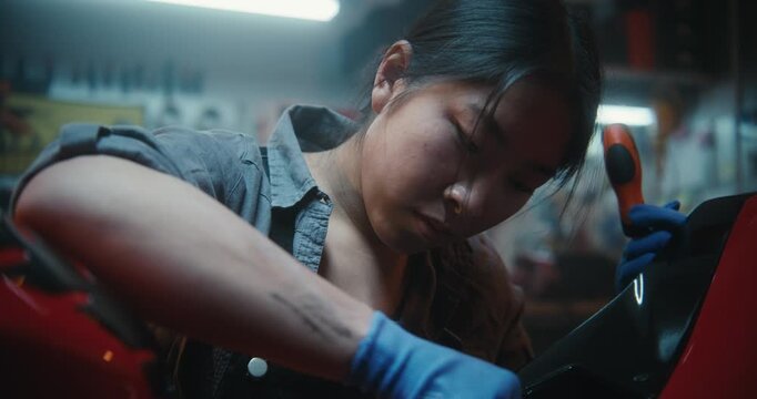 Concentrated Asian Female Mechanic Dressed in Work Gear, Fixing Motorcycle, Focusing on Repairing Electronic Components in Authentic Garage Setting. Hobbies, Female Empowerment, Automotive Culture.