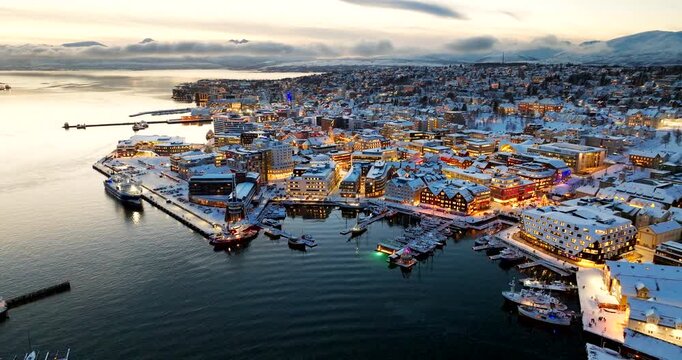 Snowy downtown Tromso city harbor and streets illuminated during twilight, drone