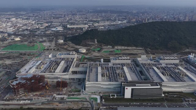 Aerial drone view of a sprawling semiconductor manufacturing complex and new construction beside an urban cityscape and green mountain, highlighting high tech industry growth
