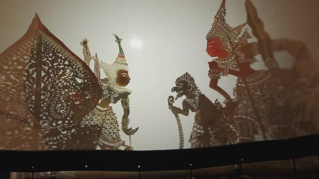 A close-up of intricate leather shadow puppets, including Rama and Hanuman characters, performing behind a backlit screen during a traditional Wayang Kulit show.
