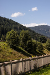 A peaceful rural scene featuring a rustic wooden fence along a grassy slope, with lush green trees and rolling forested mountains under a bright blue sky with scattered clouds.