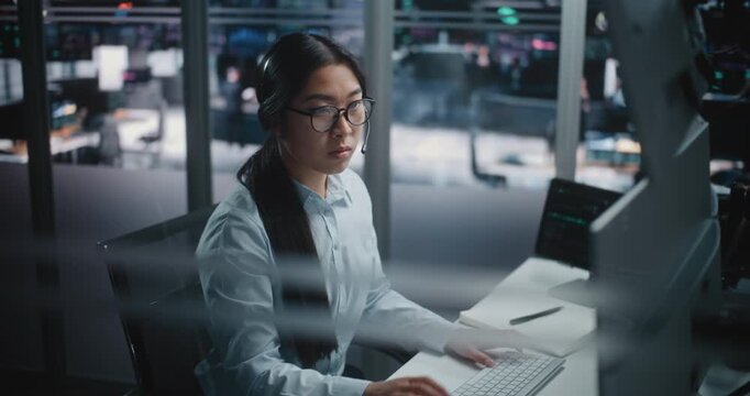 Asian Female Financial Specialist Sits at Innovative Workspace, Eyes Scanning Multiple Displays Filled With Trading Indices. Young Lady Actively Uses Keyboard to Input Data and Execute Market Orders.