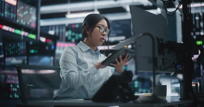 Focused Asian Female Trader Analyzing Market Trends via Tablet and Multi Monitor Setup. Woman in Glasses Sits at Workstation, Cross Referencing Real Time Data on Digital Tablet and Talking on Headset.