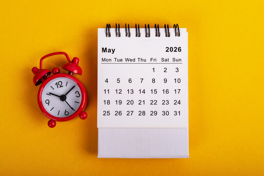 White spiral desk calendar for May 2026 placed next to a red vintage alarm clock on a solid yellow background.