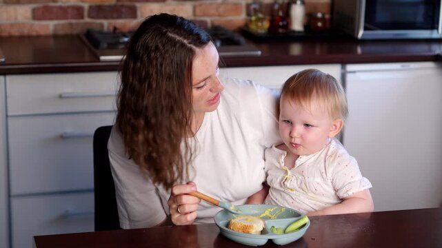 Happy mother spoon-feeds baby while sitting at table, holding him in arms and miming flight of airplane with spoon. Baby applauds as tries food for first time. Infant feeding, infant interest in food
