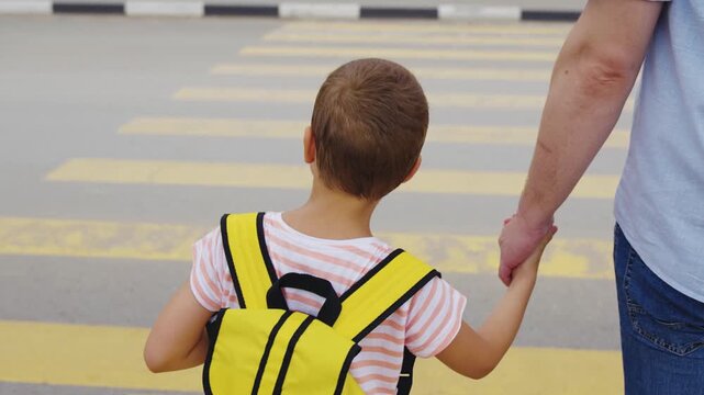 School run with father, Little boy&rsquo;s morning routine, Dad ensuring child safety, Urban crosswalk with family, Yellow backpack for visibility, Responsible parenting in action, Together on the way to