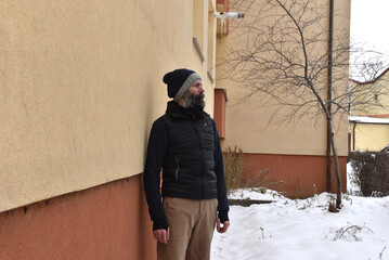 Bearded man in black gilet and pompom beanie in snow standing near apartment block © salajean