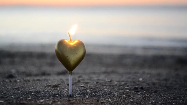 Heart shaped candle burning on sand beach near sea waves on sunset dawn. St. Valentine's Day. Burning candle in form of heart in sand against backdrop of rising sun. Romantic romance mood.