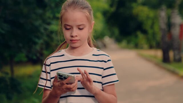 smiling child on smartphone outdoors, cute kid chatting on mobile in park, portrait of girl on phone in summer park, cute girl holding smartphone in green park, cheerful child holding phone in park