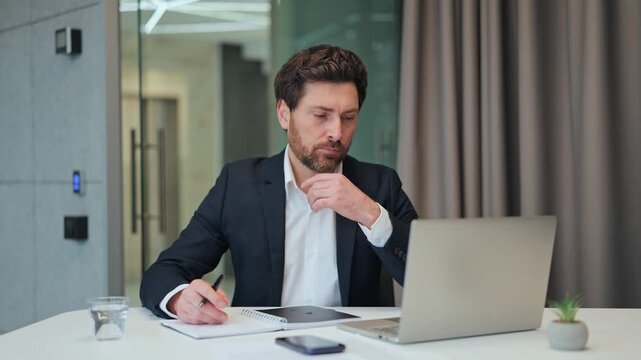 Professional man diligently working at office desk. Notebook and laptop are central to the task, showing concentrated effort and thoughtful consideration for ongoing business operations.