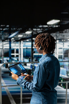 Black woman IT technician with headset and microphone does diagnostics on tablet, industrial platform. Female engineer monitoring servers infrastructure for remote troubleshooting.