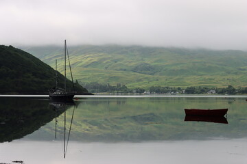 Fototapeta premium Reflections of boats and a green misty hillside on a still sea loch