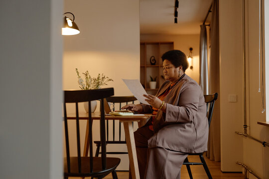 Middle aged Black woman sitting at table reviewing billing statement with focused expression, holding paper in hand, working in modern home interior, using calculator and pen, analyzing information