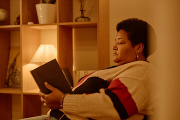 Naklejka premium Middle aged Black woman sitting indoors reading book, holding hardcover in hands, appearing focused on pages, bookshelf and lamp visible in background, relaxed posture, side profile
