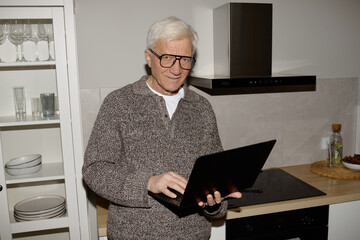 Senior Caucasian man standing in modern kitchen using laptop computer, wearing glasses and smiling...