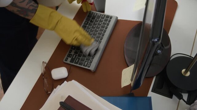Handheld high angle view of tattooed hands of unrecognizable male janitor in gloves spraying detergent and wiping computer monitor and keyboard with rag on office table