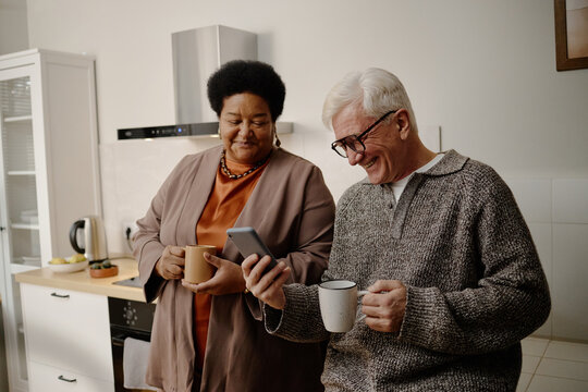 Senior Caucasian man smiling while showing smartphone to senior Black woman standing together in kitchen holding mugs, both engaging in friendly conversation and sharing moment
