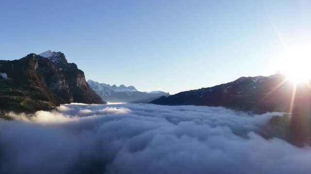 Walensee region Switzerland drone flight above thick morning clouds between Amden and Walenstadt, snow covered alps glowing in sunrise light with fog inversion landscape.