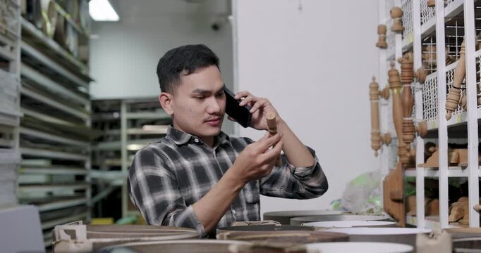 Asian adult male woodworking business owner holding wooden spindle while talking on phone, slowly turning material to confirm order details, inside interior construction material shop