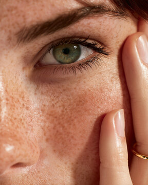 Green eye of woman with clear skin and gold ring touching cheek in skincare beauty concept on white background