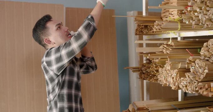 Asian adult man carpenter reaching shelf to pull bundled wood pieces, checking quantity and condition, managing inventory arrangement inside interior construction material shop for furniture projects