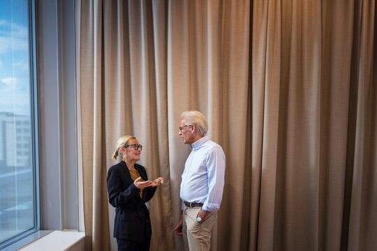 Senior businesswoman communicating with male colleague standing in front of curtain at office
