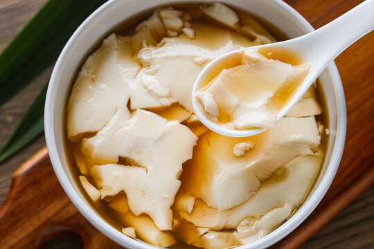 Closeup view of a delicious bowl of ginger tofu pudding (Kembang Tahu), a beloved traditional snack known for its silky texture. Captured with natural light on a wooden background.