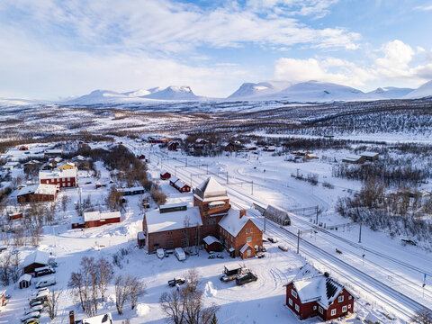 Aerial view of the Abisko train station nestled amidst a snow-covered landscape, framed by distant, majestic Lapporten mountains under a vast, open sky, Abisko, Norrbotten County, Sweden.