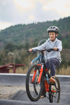 Older brother riding a mountain bike over rollers on an outdoor pump track while his younger sibling follows behind