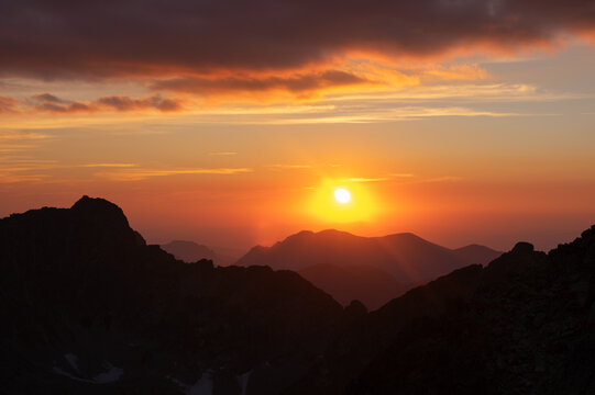 Aerial view of the sun's radiant glow paints the sky in hues of orange and gold above the dark, majestic peaks of Satan, Vysoke Tatry, Presov Region, Slovakia.
