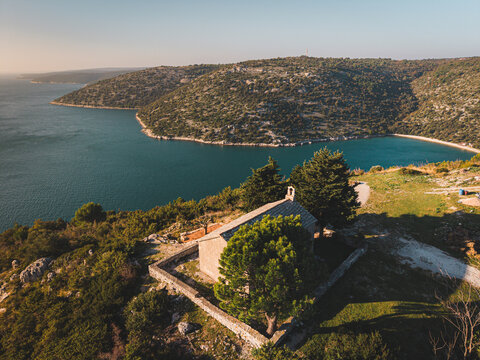 Aerial view of a quaint chapel perched atop a hill overlooking the shimmering Adriatic, framed by rugged coastline and verdant hillsides, Rakalj, Istria County, Croatia.
