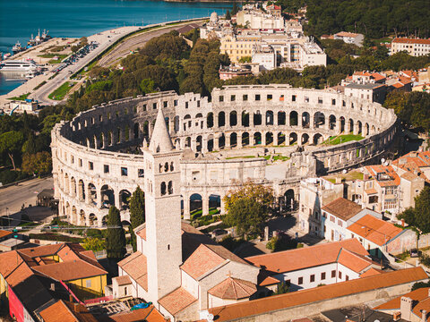 Aerial view of the majestic Pula Arena stands in ancient splendor against the backdrop of the Adriatic Sea, framed by terracotta rooftops and lush greenery, Pula, Istria County, Croatia.