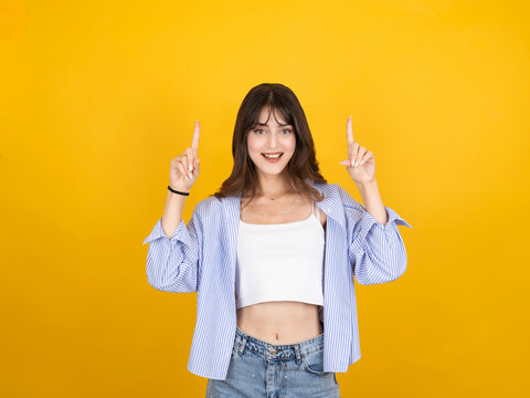 Cheerful young woman pointing fingers up at copy space, looking at camera with happy smile, isolated on yellow background. Female showing blank space for text, logo or advertisement, promotion concept