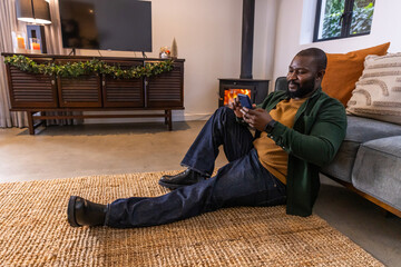 Naklejka premium African American adult man lounging on rug in living room in sweater using smartphone, copy space