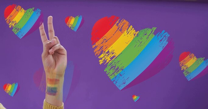 Showing arm doing V sign on purple backdrop, mustard knit sleeve, pride stripes, hearts, copy space
