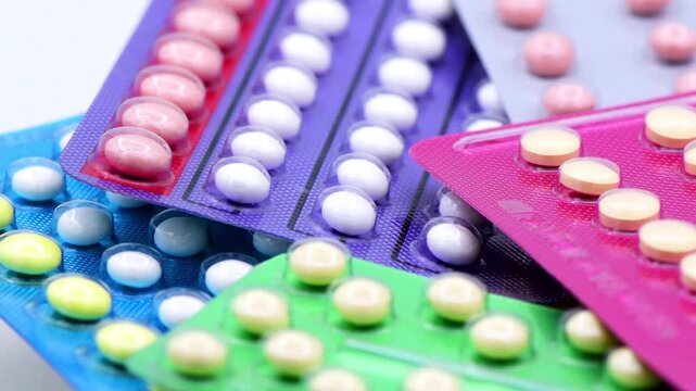 A close-up, high-angle shot showing a variety of oral contraceptive pills (COCP) arranged in a pile. The image features multiple blister packs in vibrant colors like pink, green, blue, and purple, con
