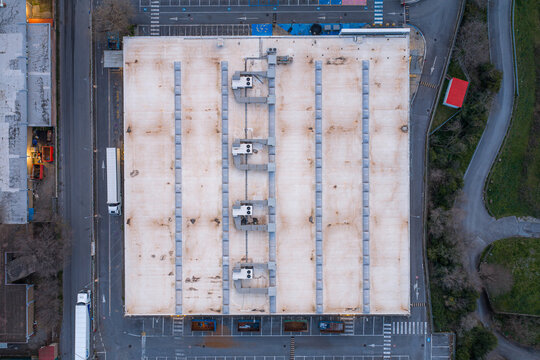 Aerial view of a large, flat rooftop with ventilation units casting shadows, bordered by a road with trucks and a winding path, Settecamini, Lazio, Italy.