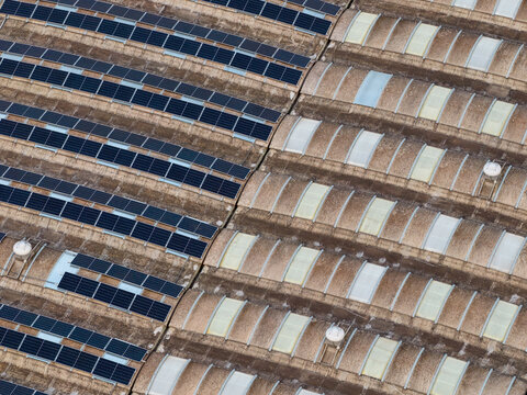 Aerial view of industrial rooftops split between dark photovoltaic panels and light, arched skylights creating a geometric contrast, Settecamini, Lazio, Italy.