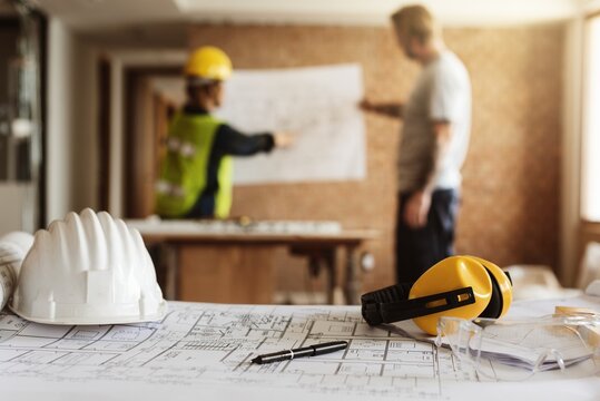 Construction site with two workers reviewing blueprints. Safety gear like helmets and headphones are visible. Focus on teamwork and planning in construction. House renovate and construction concept.